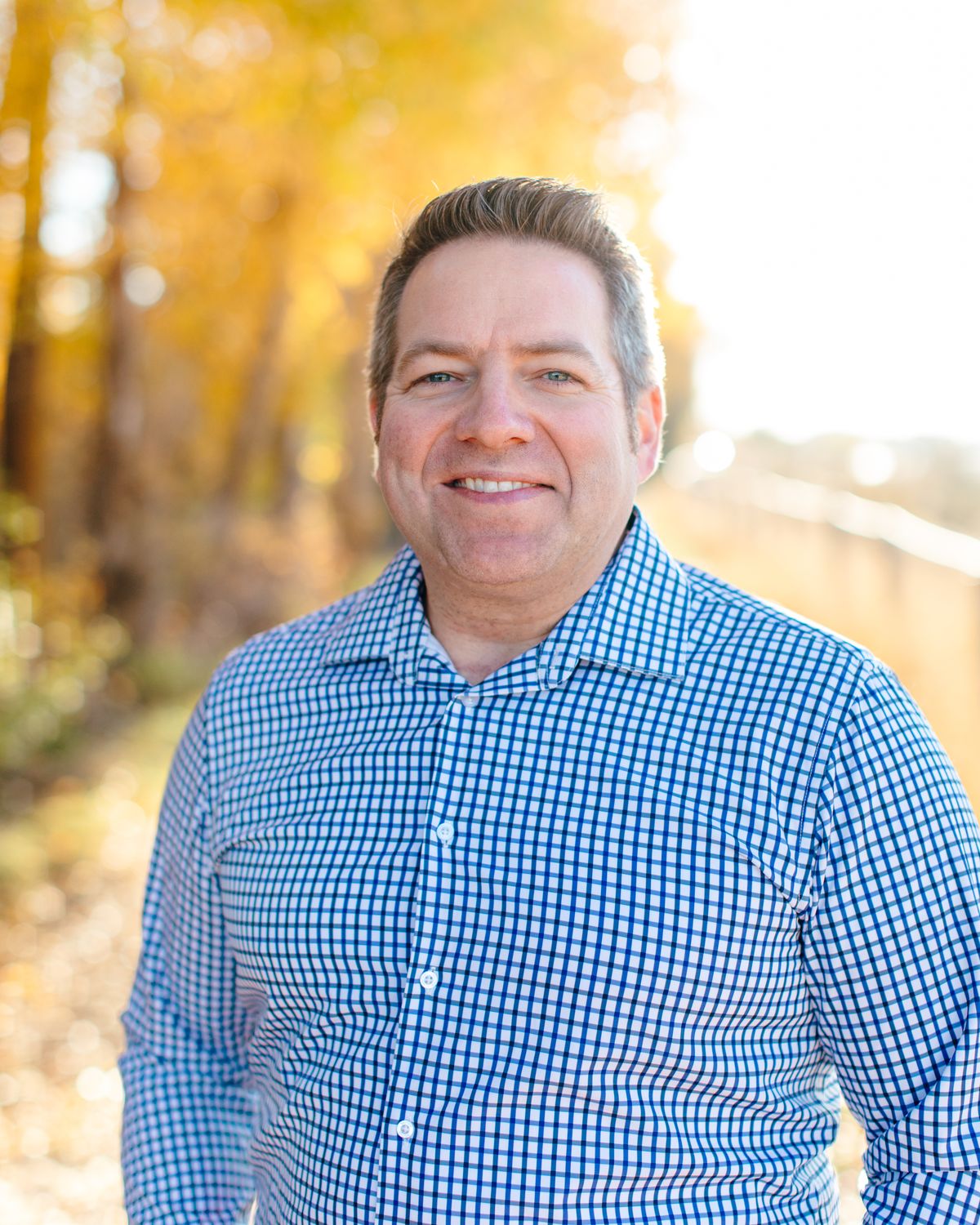 Adam Bostrom smiling outdoors in front of golden autumn trees